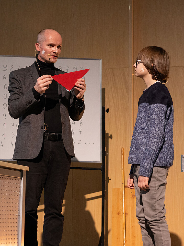 dwie osoby na tle tablicy, jedna trzyma czerwony tójkąt