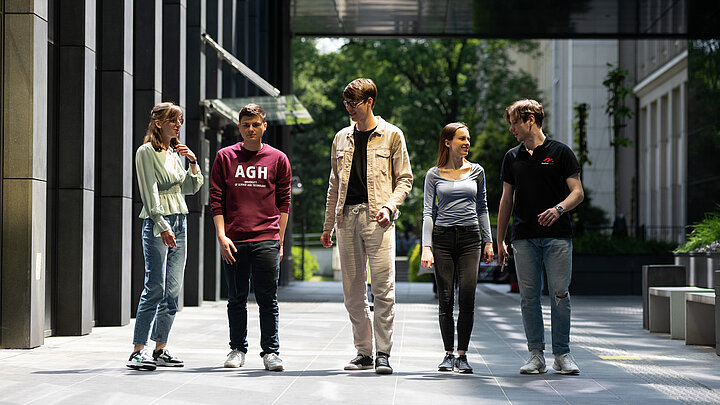 Grupa młodych ludzi pomiędzy elewacjami nowoczesnych budynków.