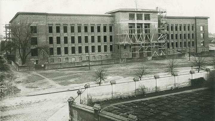 Czarno-biała fotografia przedstawiająca etap budowy dużego czterokondygnacyjnego budynku z wysuniętym do przodu portalem. Przy elewacji widoczne rusztowania.