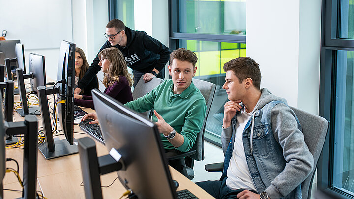 Grupa młodych ludzi pracujących przy komputerach. Na pierwszym planie dwóch mężczyzn rozmawia ze sobą.